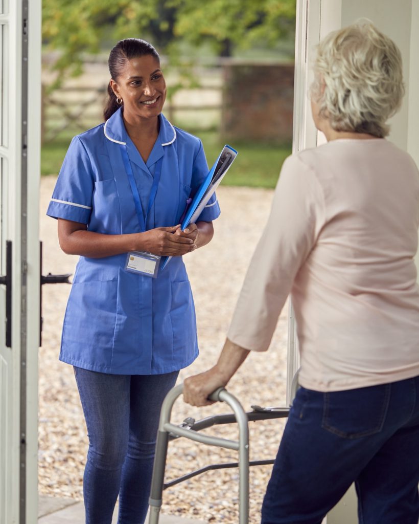 Senior Woman At Home Using Walking Stick Greeting Female Nurse Or Care Worker In Uniform At Door